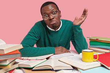 Uncertain puzzled young dark skinned man spreads palm in bewilderment, wears glasses and jumper,...