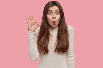 Studio portrait of shocked amazed woman makes okay gesture with suprised expression, stands over...