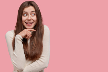 Isolated shot of dreamy beautiful woman keeps hand under chin, looks positively aside, smiles broadly, dressed in casual outfit, stands against pink studio wall with empty space for your advert