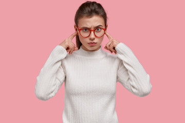 Dissatisfied young woman plugs ears with index fingers, has irritated facial expression, avoids negative sound, annoyed with loud music, dressed in white clothes, isolated over pink background