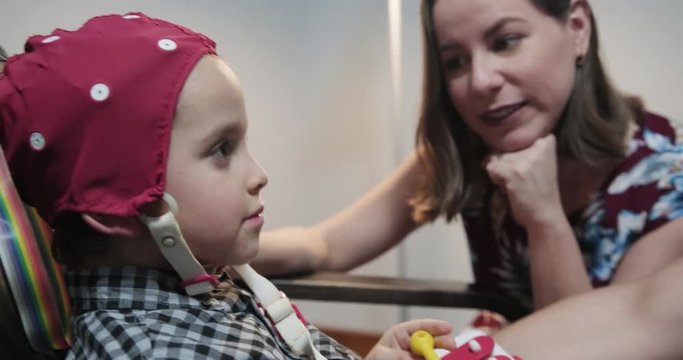 Doctor joking, performing EEG exam to child in hospital laboratory. Pediatrician doing brain therapy on boy with mom in clinic and playing with him. Lab worker using equipment on patient with mother