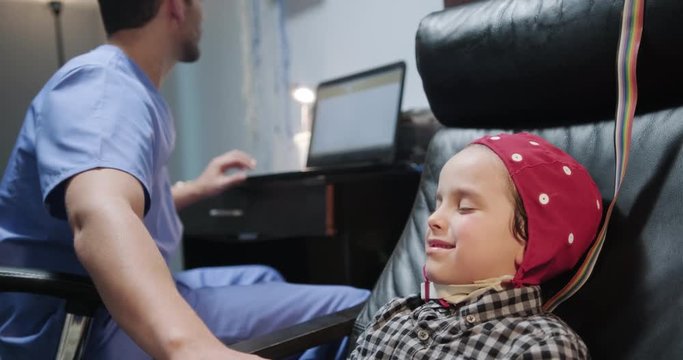 Doctor performing EEG exam to child in hospital laboratory. Pediatrician doing brain therapy on boy with mom in clinic. Lab worker using equipment on ill kid with mother. Young people and health care