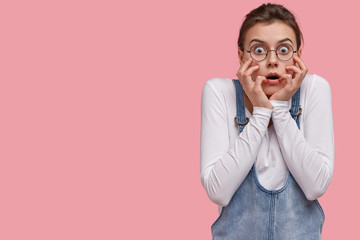 Human facial expressions and emotions concept. Woman in despair and shock, stands in stupor against pink background, touches face, has scared look, wears round spectacles and denim overalls.