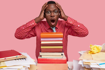 Photo of astonished black man keeps hands on head, stares with overwhelmed expression, has to read all books for exam, prepares annual report, dressed in formal clothes, wears square spectacles