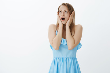 Waist-up shot of shocked and upset charming young woman with fair hair in blue dress dropping jaw being concerned and troubled holding hands on cheeks looking perplexed at upper left corner