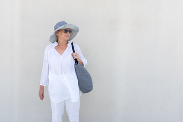 Beautiful senior woman in grey hat and light white summer clothing against wall (selective focus)
