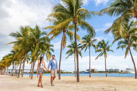 Miami people lifestyle - couple walking holding hands talking enjoying walk on beach with palm trees. Florida travel destination.