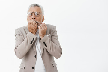 Old rich man scared losing money. Portrait of nervous afraid senior male model in glasses and suit biting fingers from anxiety and fear panicking staring terrified at camera against white background