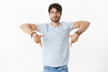 Portrait of handsome charming young european male with big blue eyes and bristle pointing down indicating at copy space, smiling friendly and assured, advertising place to put promotions