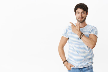 Fototapeta premium Skillful male entrepreneur showing product to potential customer standing confident and relaxed pointing at upper left corner holding hand on waist and smiling self-assured over white background
