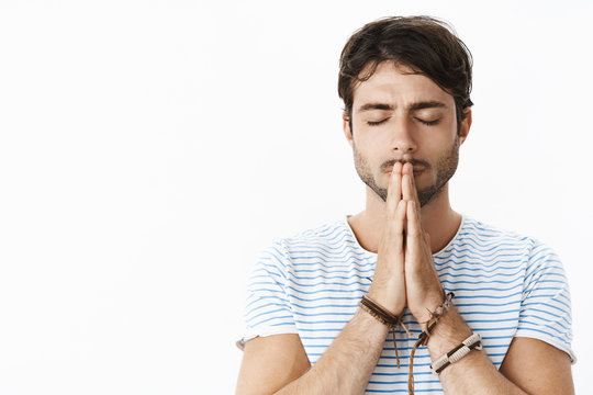 Focused young hopeful hispanic guy with beard in striped t-shirt holding hands in pray over lips standing with closed eyes as praying, talking to god, making wish, anticipating for miracle happen