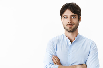 Waist-up shot of successful delighted good-looking young male with blue eyes and beard smiling satisfied as holding hands crossed on chest being self-satisfied achieving good income over grey wall