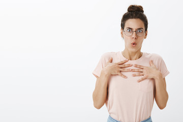 Offended and shocked female tutor in glasses and hair bun holding breath and hands on chest in...