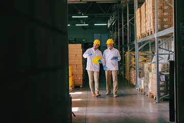 Two workers with helmets on heads walking in warehouse. All around shelves and boxes.