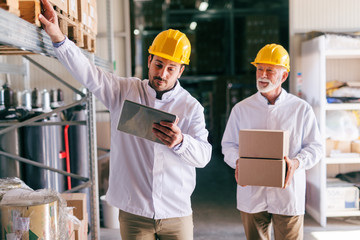 Young male Caucasian worker using tablet while standing in warehouse. In background other worker...
