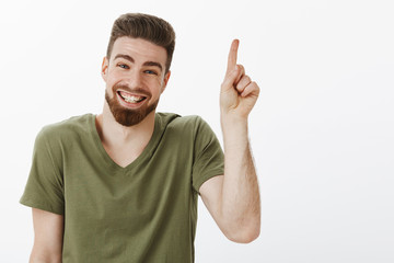 Fototapeta premium Happy delighted and carefree attractive adult man with beard laughing joyfully having great time looking delighted and joyful at camera pointing up or showing number one over white wall
