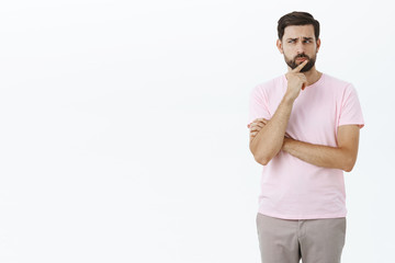 Portrait of creative handsome adult bearded male in pink t-shirt making hmm sound holding finger on lips frowning looking determined and thoughtful gazing left while thinking over gray wall