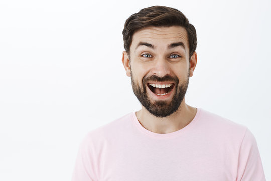 Headshot of amused excited joyful 30s man with beard laughing out loud, having fun standing entertained and amazed over gray wall giggling joyfully raising eyebrows as chuckling over hilarious joke