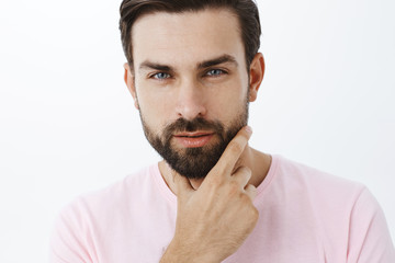 Headshot of handsome cheeky and sensual sexy european man with beard and blue eyes squinting with desire and seduction gaze touching jawline, looking at camera confident and self-assured
