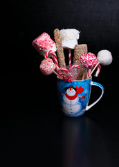Blue Christmas mug with a snowman filled with candy pops, marshmellows and candy canes on a black background with text space.  Christmas baked gifts.
