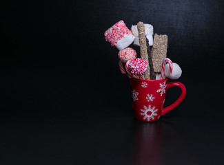 Red Christmas mug with snowflakes filled with cake pops, candy canes and marshmallows on a black background with text space. Christmas baked gifts.