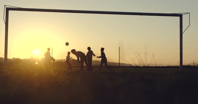 View from the below and grass on the teens playing football village match and running with a ball on the sunset. Outdoors.