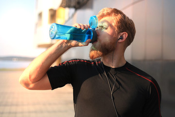 Handsome adult man drinking water from fitness bottle while standing outside, at sunset or sunrise. Runner.