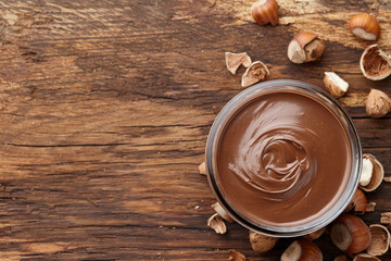 Chocolate spread or nougat cream with hazelnuts in glass jar on wooden background, top view