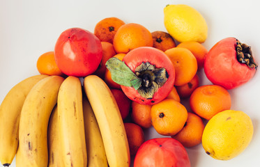 A selection of arranged different fresh fruits of bananas, mandarins, persimmons and lemons on white background close up.