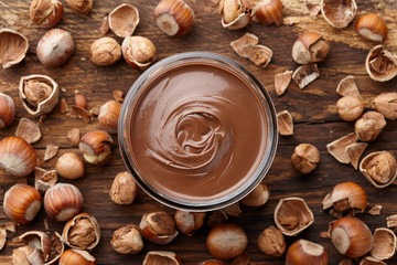 Chocolate spread or nougat cream with hazelnuts in glass jar on wooden background, top view