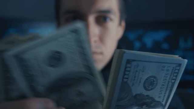 Portrait of Hacker Programmer counts Money earned ith criminally on Darknet in cyber security center filled with display screens.