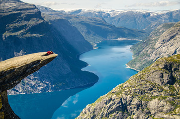 A man l in a red jacket aying on the Trolltunga rock with a blue lake 700 meters lower and interesting sky with clouds