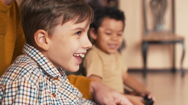 Diverse boys sitting near on the floor at home and playing video games with the help of their fathers who holding on joysticks