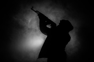 Silhouette of man with assault rifle ready to attack on dark toned foggy background or dangerous bandit in black wearing balaclava and holding gun in hand. Shooting terrorist with weapon theme decor - obrazy, fototapety, plakaty