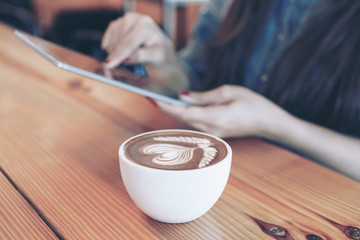 Fototapeta na wymiar white coffee cup with latte art heart pattern on a wooden table background and Young woman hand holding smartphone in a coffee shop cafe