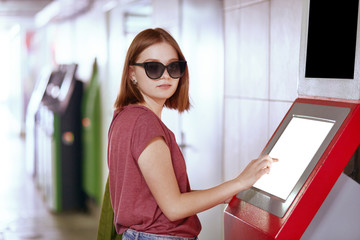 Fototapeta na wymiar Shot of pretty Caucasian woman in trendy sunglasses, poses near ATM machine with blank screen, needs to withdraw money, stands in underground, carries green rucksack. People and payment concept