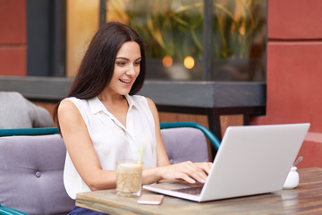 Positive dark haired woman in elegant clothes types on laptop computer, chats with friends, browses...