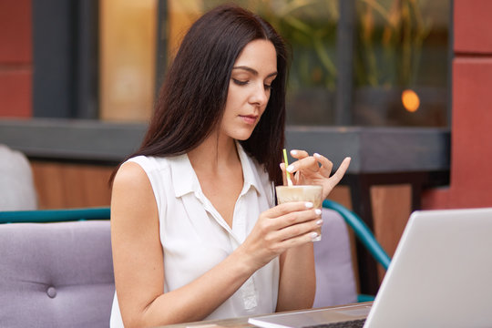 Beautiful brunette female student works on scientific project via laptop computer, searches online, dressed in elegant white blouse, hold glass of cocktail, spends free time in outdoor cafeteria.