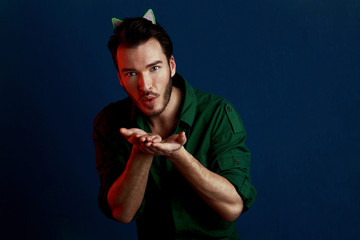 Lovely man in cat ears on the head rounds lips as blows air kiss, tries to flirt, man in green shirt and style haircut poses over dark blue wall with red contrast color light