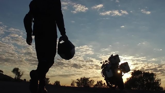 Silhouetted motorcyclist walking with helmet towards his motorbike at sunset