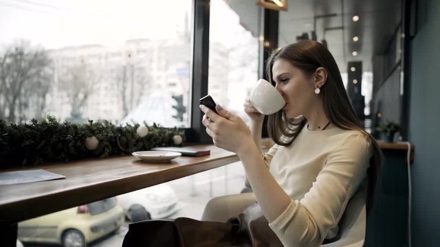 A young girl drinks coffee and uses a smartphone. Woman holding a cup of coffee. Business lady sits in a cafe with a phone and coffee