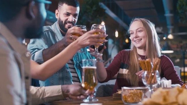 Over-the-shoulder shot of couple coming closer to their friends sitting at the table in bar, greeting them and clinking glasses with beer