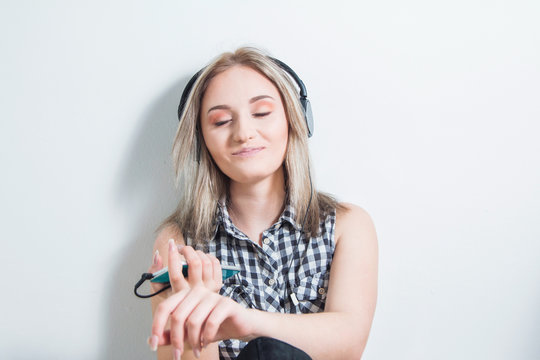 Happy young girl in plaid shirt with headphones and smart phone sitting at floor, smiling and enjoying music at home. White wall, half body, youth music concept. 
