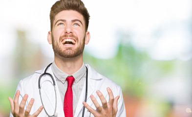 Young handsome doctor man wearing medical coat crazy and mad shouting and yelling with aggressive expression and arms raised. Frustration concept.