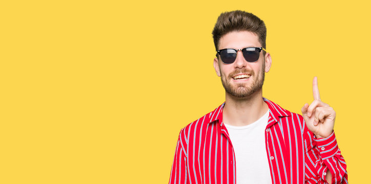 Young handsome man wearing sunglasses pointing finger up with successful idea. Exited and happy. Number one.