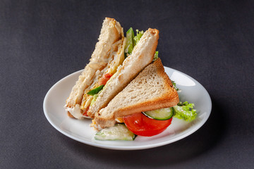 Homemade sandwich made of toast bread, fried egg, melted cheese, tomatoes, cucumber, lettuce, burgers and mayonnaise sauce. Sandwich on a white plate, on a black background.