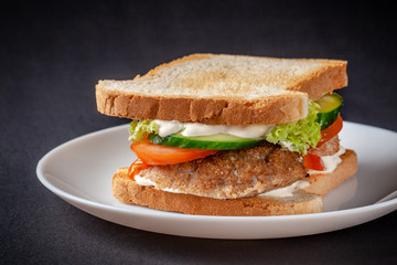 Homemade sandwich made of toast bread, fried egg, melted cheese, tomatoes, cucumber, lettuce, burgers and mayonnaise sauce. Sandwich on a white plate, on a black background.