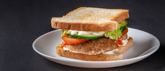 Homemade sandwich made of toast bread, fried egg, melted cheese, tomatoes, cucumber, lettuce, burgers and mayonnaise sauce. Sandwich on a white plate, on a black background.