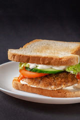 Homemade sandwich made of toast bread, fried egg, melted cheese, tomatoes, cucumber, lettuce, burgers and mayonnaise sauce. Sandwich on a white plate, on a black background.