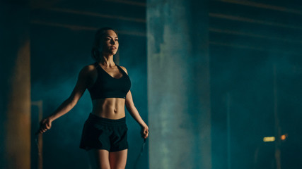 Fototapeta na wymiar Beautiful Energetic Fitness Girl in Black Athletic Top and Shorts is Skipping/Jumping Rope. She is Doing a Workout in an Evening Foggy Urban Environment Under a Bridge.